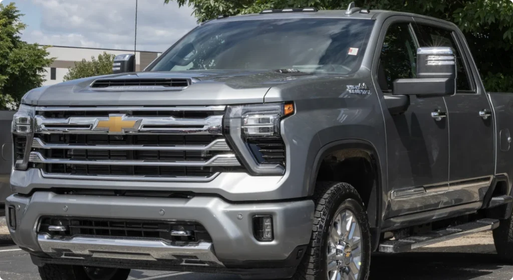 A silver Chevrolet Silverado HD pickup truck is parked outdoors, showcasing its large front grille, chrome accents, and side mirrors, with trees and buildings in the background.