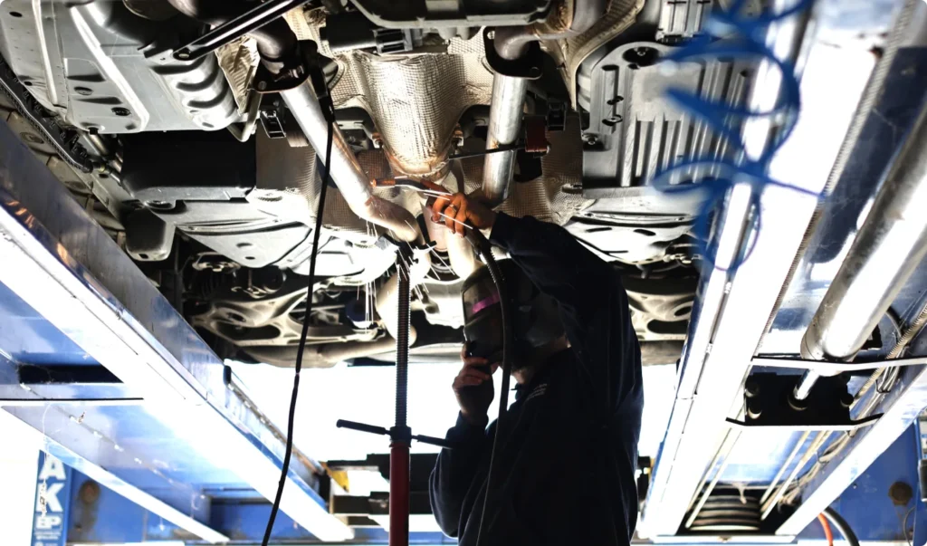 Technician welding a vehicle’s exhaust system while elevated on a lift, illustrating complete auto repair and undercar service in a professional shop setting.