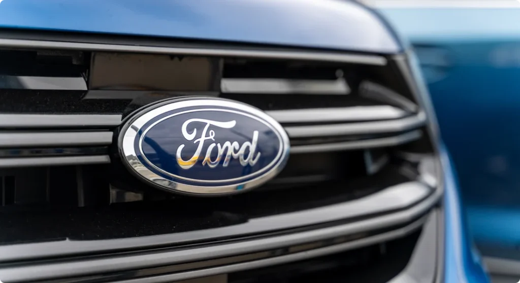 Close-up of a blue Ford vehicle’s front grille, featuring the Ford logo prominently in the center, with another blue vehicle blurred in the background.