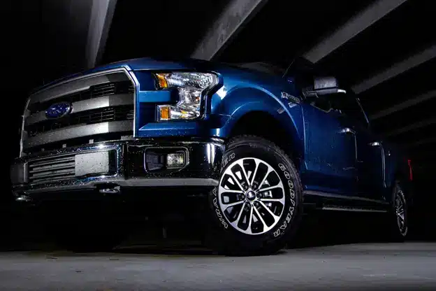 Blue Ford F-150 pickup truck with chrome accents parked under an overpass, viewed from a low angle highlighting the front grille and headlights.